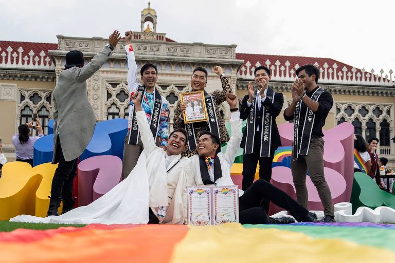 Um casal recém-casado sorri radiante, sentado em frente a uma bandeira do orgulho LGBTQIA+ colorida, enquanto um grupo de amigos os felicita com entusiasmo. O cenário é adornado com uma arquitetura elegante ao fundo, e um dos amigos segura um cinturão comemorativo, simbolizando a vitória e a celebração do amor.