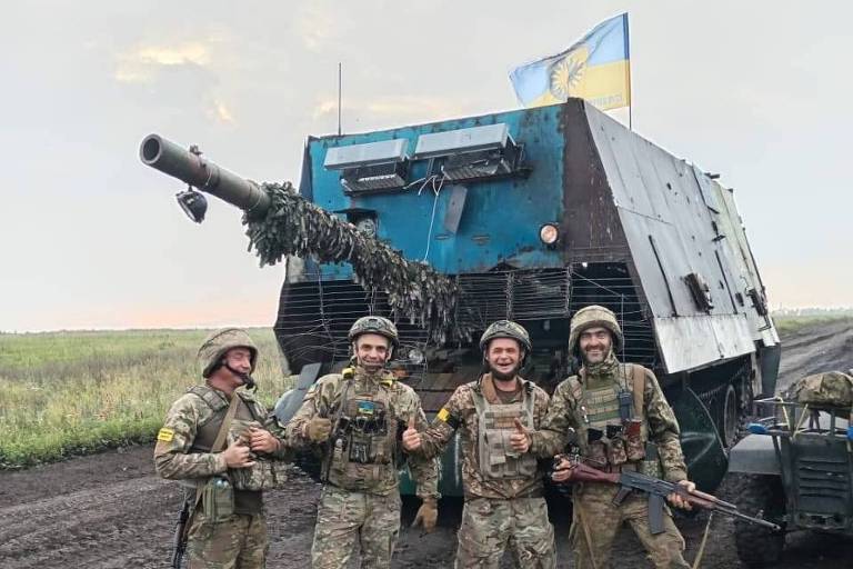 Quatro soldados uniformizados à frente de um tanque com uma cobertura de metal azul, com uma bandeira da Ucrânia em cima