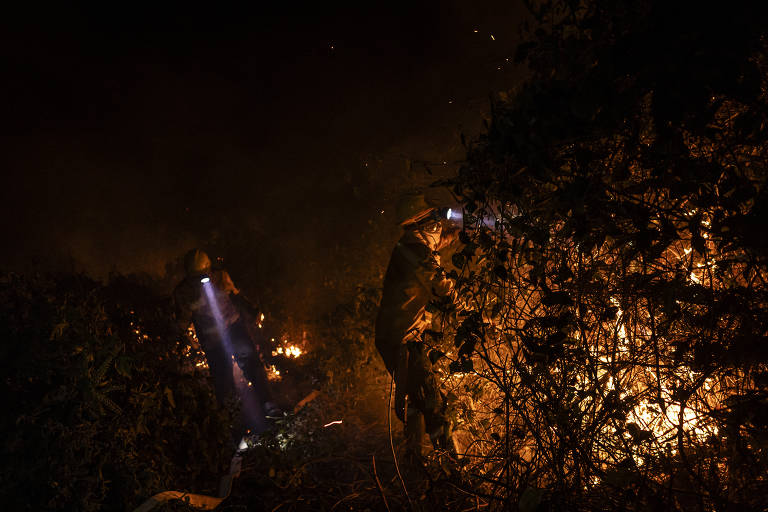 foto à noite mostra dois brigadistas, equipados com capacetes com lanterna e uniformes, enfrentando fogo. eles são iluminados pelas chamas