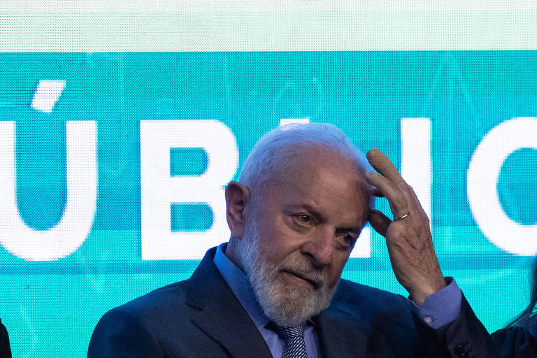 Lula, um homem idoso com cabelo e barba grisalhas, está com expressão pensativa e toca sua cabeça, enquanto está em frente a um fundo azul com a palavra "PÚBLICO" parcialmente visível. A postura contemplativa sugerem um momento de reflexão ou uma pausa em um evento público
