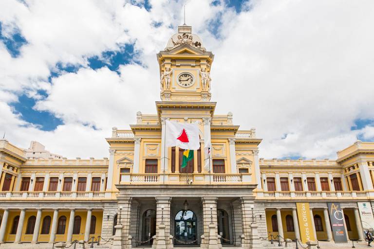 A fachada imponente do edifício histórico exibe uma arquitetura neoclássica, com um relógio no topo e bandeiras tremulando ao vento, sob um céu parcialmente nublado que realça a grandiosidade do local.