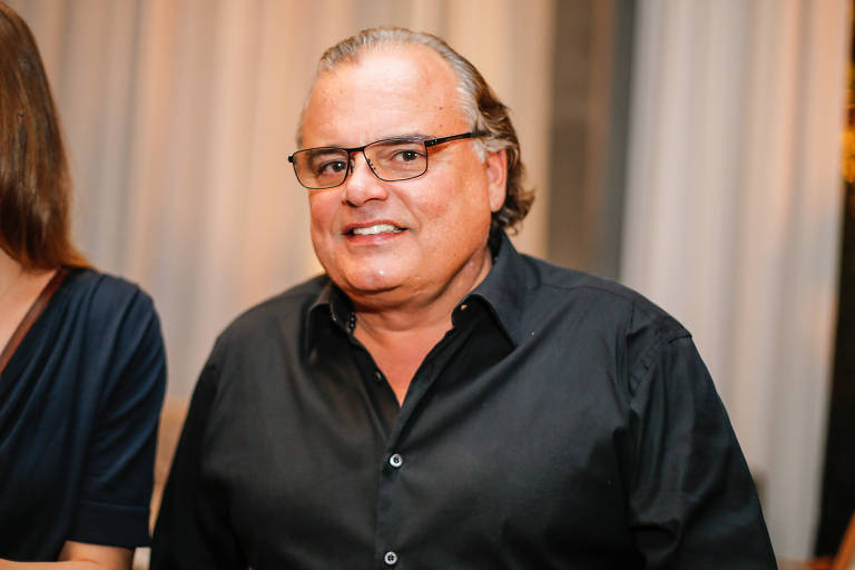 Homem de óculos, cabelo penteado para trás e camisa social preta posa com sorriso para foto