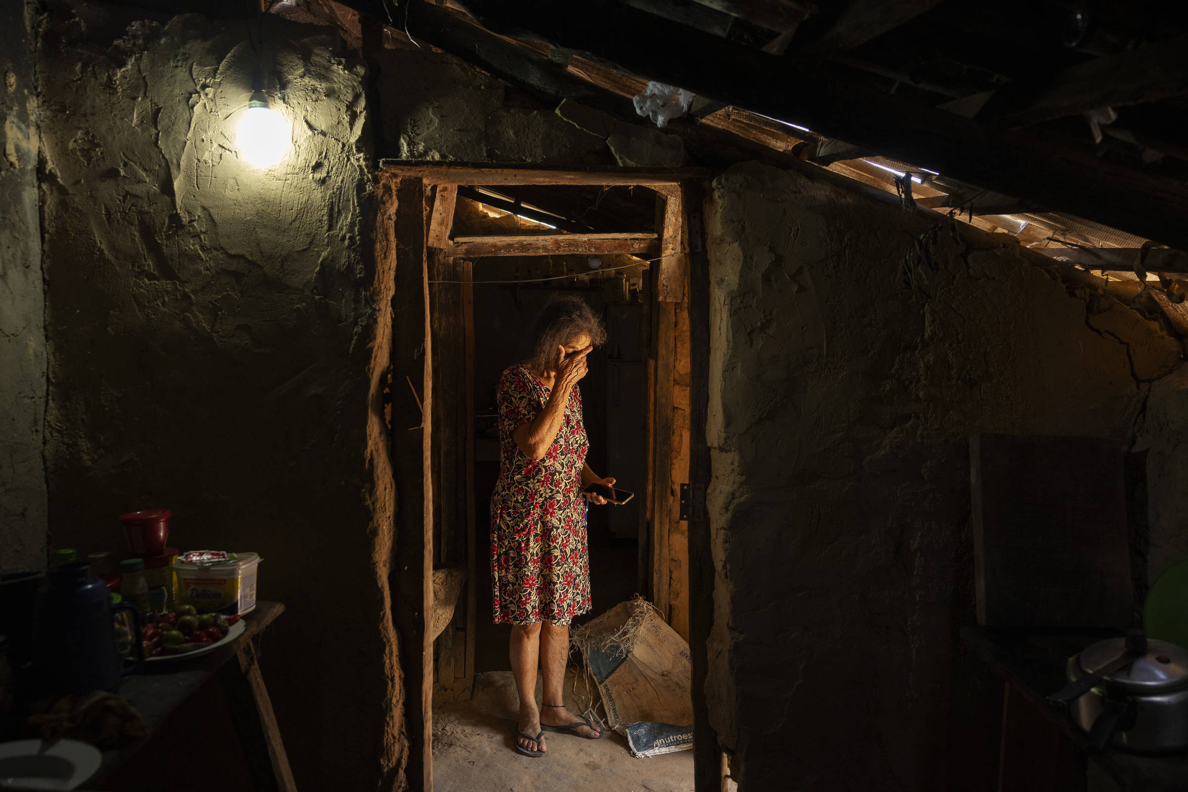 Uma mulher em um vestido estampado apoia a cabeça nas mãos, parada na entrada de uma casa rústica, com paredes de barro