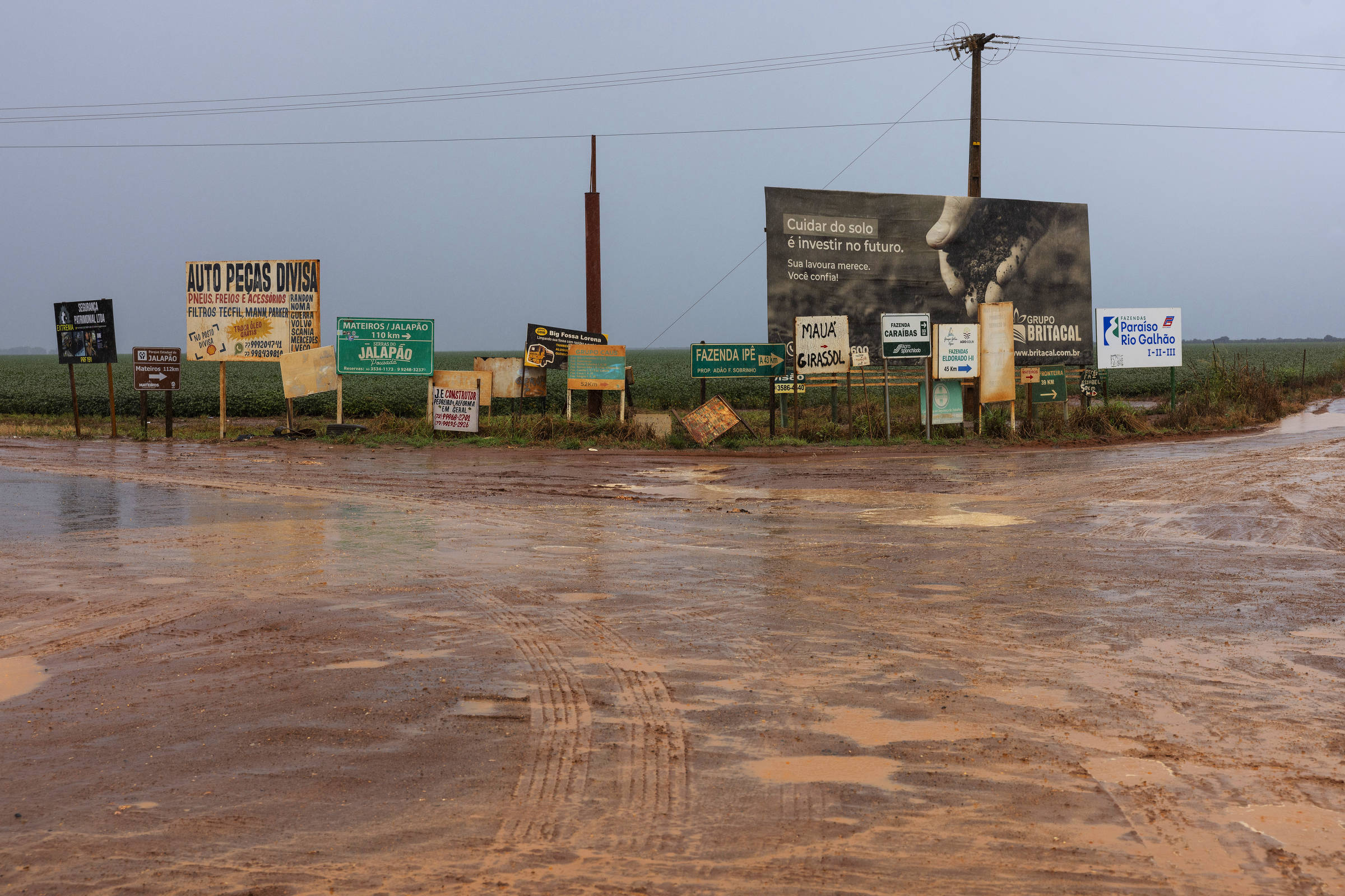 Uma estrada de terra molhada se estende até o horizonte, ladeada por uma variedade de placas de propaganda e sinalização