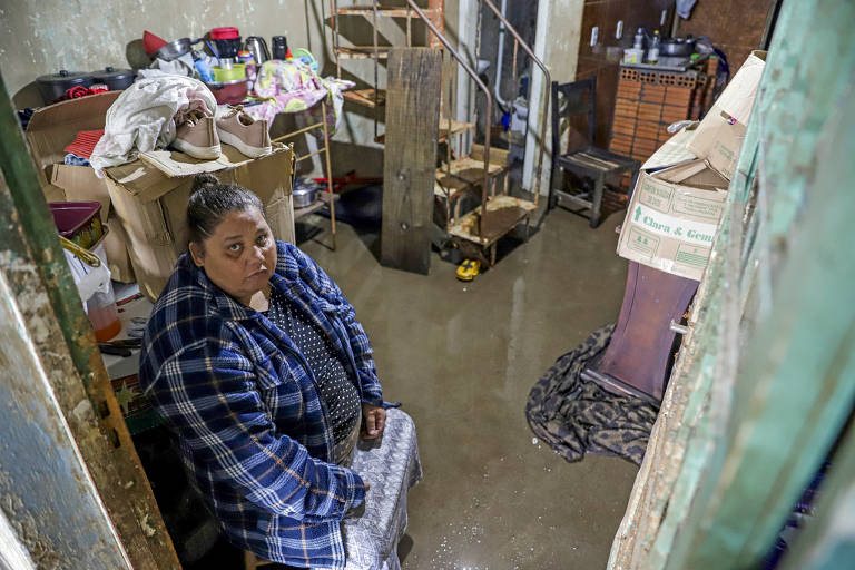 Uma mulher sentada em uma cadeira olha para cima com uma expressão de preocupação e resiliência, enquanto água inunda o chão de sua casa, cercada por pertences empilhados de forma precária para evitar danos