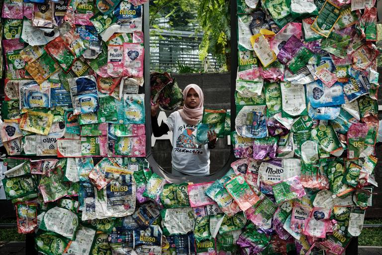 A imagem mostra uma mulher usando um hijab branco e uma camiseta branca, em pé atrás de uma moldura feita de várias embalagens recicladas de produtos diversos. As embalagens são coloridas e cobrem toda a moldura, exceto por uma abertura retangular no centro, onde a mulher está posicionada. Ao fundo, é possível ver árvores e uma cerca.
