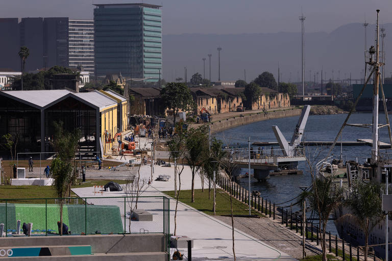 A imagem mostra um calçadão à beira-mar com várias árvores e áreas de lazer. À esquerda, há edifícios modernos e estruturas industriais. À direita, há um corpo d'água com barcos atracados e uma ponte pedonal. Ao fundo, é possível ver mais edifícios e montanhas sob um céu claro.