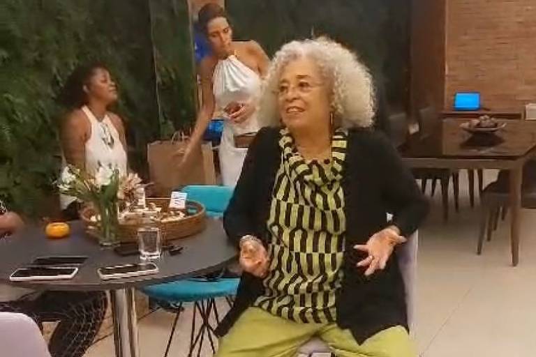 A imagem mostra uma mulher idosa de cabelos grisalhos e volumosos, vestindo uma blusa listrada em preto e amarelo e calças verdes, sentada em uma cadeira alta. Ela parece estar conversando e gesticulando. Ao fundo, há outras pessoas, incluindo uma mulher de vestido branco e outra sentada à mesa. 