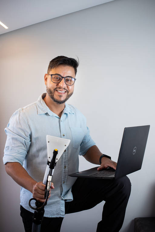 Homem branco de camisa social, óculos e barba. Ele está com um pé apoiado sobre uma superfície acima do chão. Em uma mão, segura um aspirador de pó. Na outra, um laptop