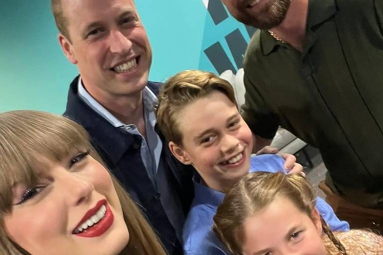 A imagem mostra cinco pessoas sorrindo para uma selfie. No canto inferior esquerdo, uma mulher com cabelo loiro e batom vermelho está vestindo uma blusa preta com detalhes brilhantes. Ao lado dela, um homem com uma camisa azul clara e um casaco escuro sorri para a câmera. No centro, duas crianças, um menino e uma menina, também sorriem. O menino está vestindo uma camisa azul clara, e a menina tem cabelo loiro trançado e veste uma blusa clara. No canto superior direito, um homem com barba e boné branco com o logo 'USA' está vestindo uma camisa polo verde escura. O fundo é azul com letras brancas e pretas parcialmente visíveis.