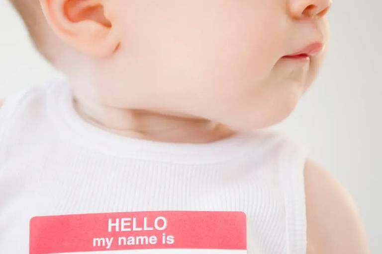 A imagem mostra um bebê vestindo uma camiseta branca sem mangas. No peito do bebê, há uma etiqueta de nome vermelha e branca com o texto 'HELLO my name is' (olá, meu nome é), mas o espaço para o nome está em branco.