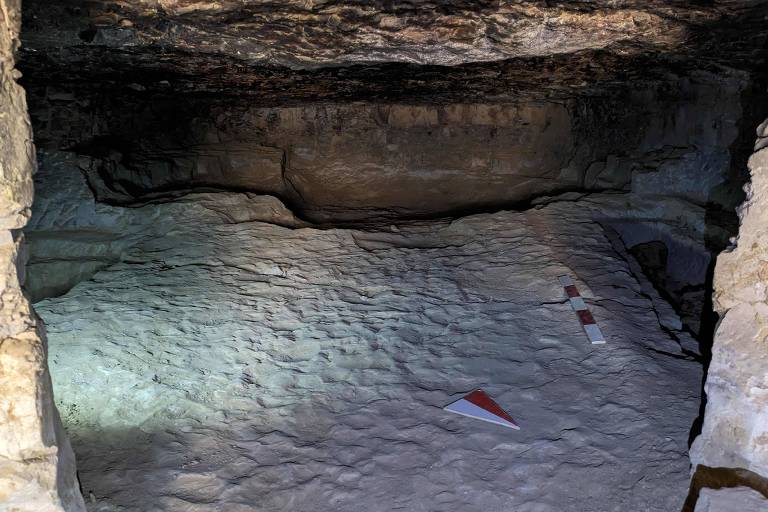 A imagem mostra o interior de uma área com um chão rochoso e irregular. Há dois marcadores de pesquisa no chão, um em forma de seta e outro em forma de régua com listras vermelhas e brancas. A iluminação é fraca, destacando a textura das paredes e do chão
