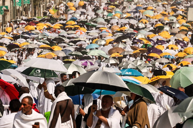 A imagem mostra uma grande multidão de pessoas vestidas com roupas brancas, muitas delas segurando guarda-chuvas coloridos. A cena parece ocorrer em uma área urbana, com sinais de trânsito visíveis ao fundo. A maioria dos guarda-chuvas são de cores variadas, incluindo amarelo, verde, azul e branco.