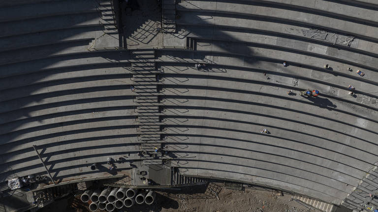 Imagem aérea de um anfiteatro em construção. A estrutura é composta por várias fileiras de assentos em concreto, dispostas em semicírculo. Há algumas pessoas trabalhando na construção, visíveis como pequenos pontos na imagem. Tubos de concreto estão empilhados no canto inferior esquerdo da imagem.