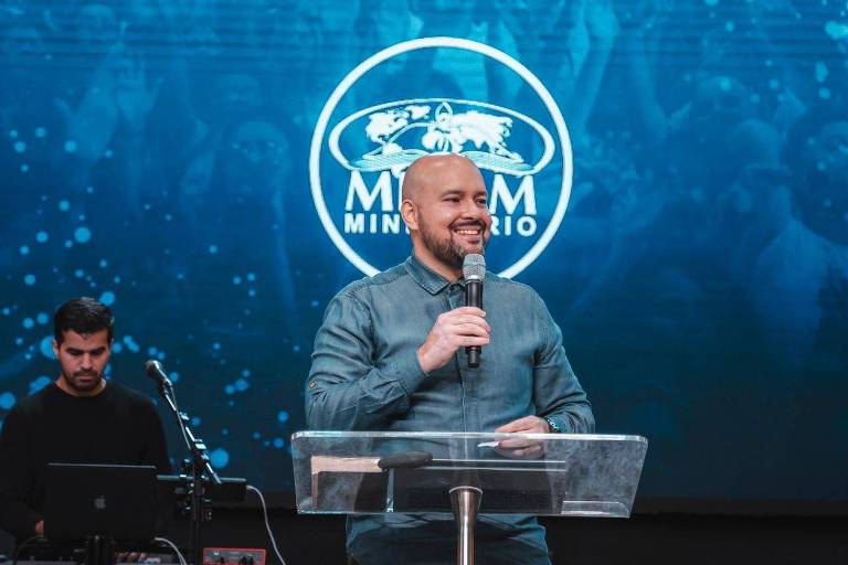 Foto colorida do pastor Roberto Oliveira pregando no Mevam. Ele é branco, careca, usa barba escura e veste camisa social azulada de manga comprida. Esté sorrindo, segurando o microfone. Ao fundo, à sua esquerda, há um tecladista de pele morena e cabelo escuro