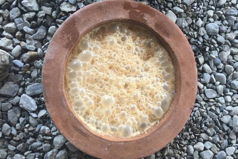 A imagem mostra uma tigela de barro cheia de um vinho de cor amarelada, que está espumando. A tigela está colocada sobre um chão coberto de pedras pequenas e irregulares.