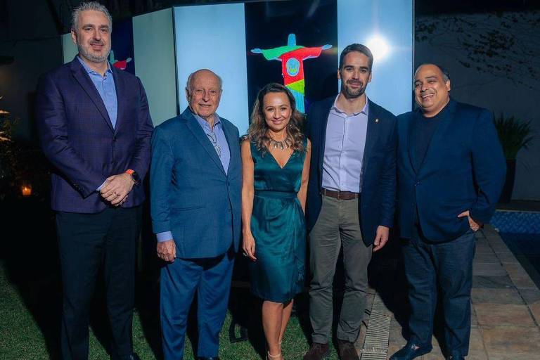 Cinco pessoas posam para uma foto em um evento. Ao fundo, há uma tela exibindo uma imagem do Cristo Redentor com um fundo colorido. As pessoas estão bem vestidas, com trajes formais. O ambiente parece ser ao ar livre, com grama e uma área pavimentada visíveis.