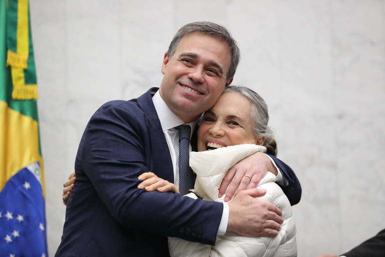 André Mendonça, de terno azul e gravata, está abraçando Regina Duarte, que veste um casaco branco. Ambos estão sorrindo. Ao fundo, há uma bandeira do Brasil parcialmente visível.