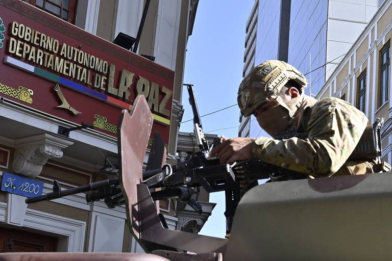 A imagem mostra um soldado em uniforme militar e capacete, manuseando uma metralhadora montada em um veículo militar. Ao fundo, há um prédio com uma placa que diz 'GOBIERNO AUTONOMO DEPARTAMENTAL DE LA PAZ'.