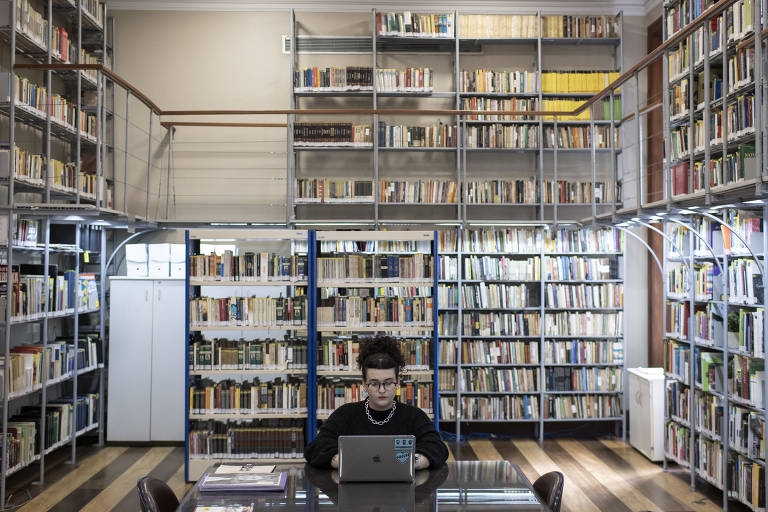 Na imagem, uma mulher de roupa preta está numa biblioteca cercada de livros, sentada à mesa com um computador.