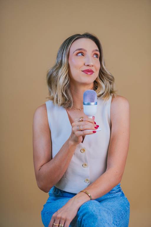 A imagem mostra uma mulher de cabelos loiros e ondulados, vestindo uma blusa sem mangas branca com botões e calça jeans azul. Ela está segurando um microfone branco