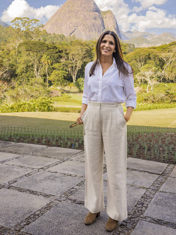 Uma mulher está de pé em uma área pavimentada ao ar livre, com uma montanha ao fundo. Ela está vestindo uma camisa branca de mangas compridas e calças largas de cor clara, com as mãos nos bolsos. Ela segura um par de óculos de sol em uma das mãos. Atrás dela, há uma área gramada e várias árvores, com um céu azul e algumas nuvens brancas