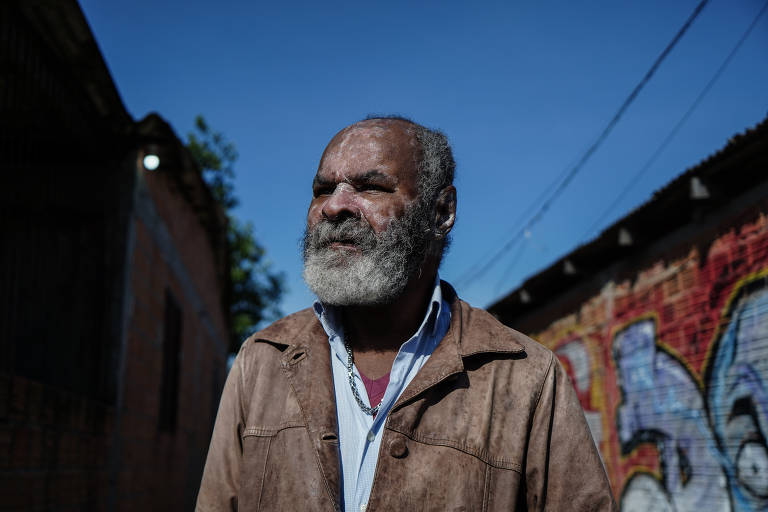 Silva é negro, tem barba branca, usa camisa azul e jaqueta marrom; o rosto tem marcas claras que parecem terra ou poeira