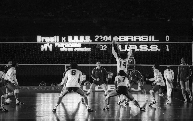 Imagem em preto e branco de uma partida de vôlei entre as seleções do Brasil e da URSS no estádio do Maracanã. O placar eletrônico ao fundo mostra 'Brasil x U.R.S.S.' com o tempo de 22:04 e o placar de 0 para o Brasil e 1 para a URSS. Jogadores de ambas as equipes estão em ação na quadra, com um jogador da URSS (número 14) saltando para atacar a bola.