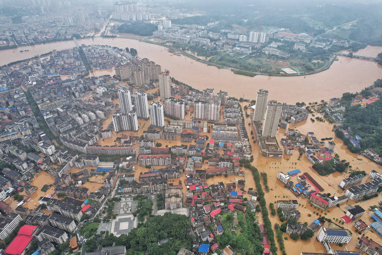 Imagem aérea de uma cidade inundada, onde várias áreas estão submersas em água barrenta. Edifícios altos e estruturas residenciais são visíveis, com ruas e terrenos cobertos por água. Um rio largo e marrom atravessa a cidade, contribuindo para a inundação. A paisagem ao fundo mostra áreas urbanas e vegetação.