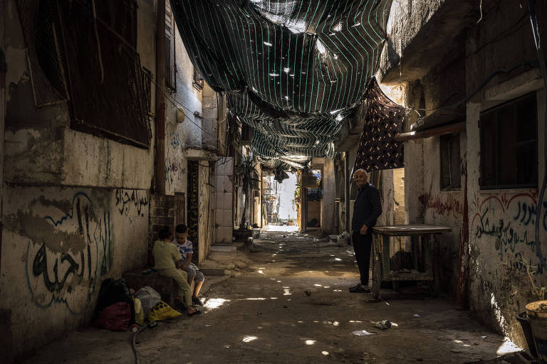 A imagem mostra um beco estreito com paredes desgastadas e grafitadas. Há uma cobertura improvisada feita de lonas e tecidos pendurados acima, permitindo a passagem de alguns raios de luz. À esquerda, duas crianças estão sentadas no chão, próximas a sacos e objetos. À direita, um homem está em pé ao lado de uma mesa de metal. O ambiente parece ser uma área urbana degradada.