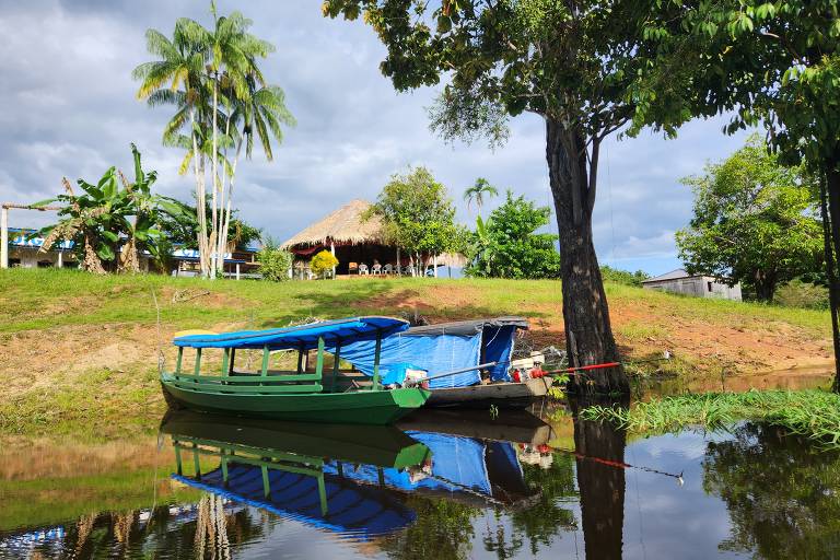 A imagem mostra dois barcos ancorados em um rio calmo, com suas reflexões visíveis na água. Os barcos têm coberturas azuis e verdes. Ao fundo, há uma cabana com telhado de palha, cercada por vegetação tropical, incluindo palmeiras e outras árvores. O céu está parcialmente nublado.