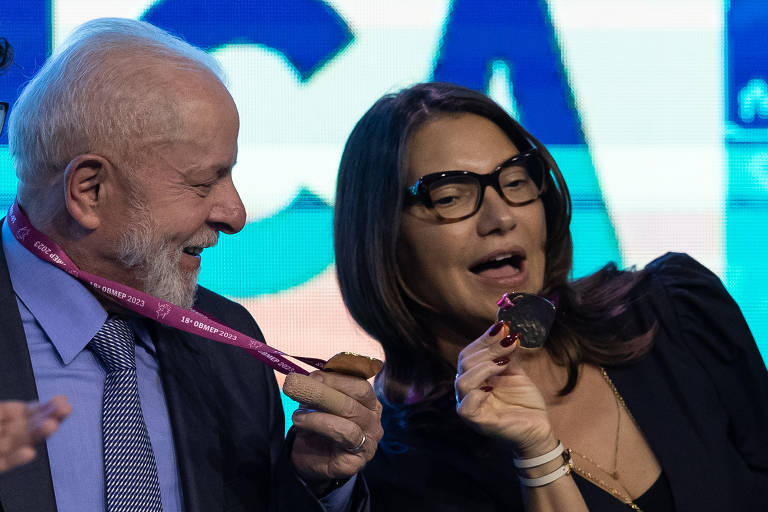 Lula, homem branco de cabelo grisalho, usa camisa social azul, paletó escuro e gravata, e a sua direita na foto, está Janja, mulher branca, de roupa preta e óculos. Lula está com o que parece ser uma medalha no pescoço e Janja a segura.