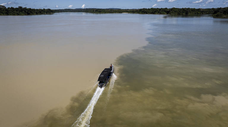 Imagem aérea de um barco navegando em um rio onde as águas de diferentes cores se encontram. O barco, com um toldo preto, está se movendo em uma área de água mais clara, enquanto a água mais escura está à direita. Ao fundo, há uma densa floresta verde e um céu azul com algumas nuvens brancas.