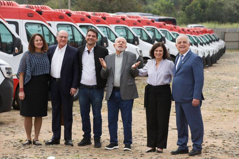 Seis pessoas estão posando para uma foto em frente a uma fila de veículos brancos com detalhes em vermelho. O grupo está em um terreno de terra e cascalho, e ao fundo há uma vegetação esverdeada. As pessoas estão sorrindo e parecem estar em um evento oficial ou cerimônia de entrega de veículos.