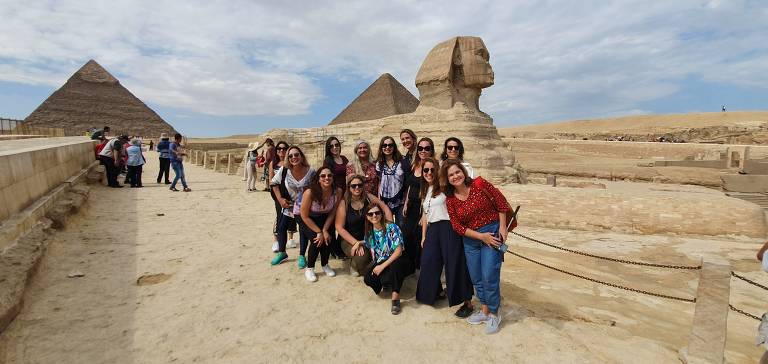 Fotografia de mulheres posando no Egito. Ao fundo, é possível ver duas das pirâmides. O céu está parcialmente nublado e o ambiente é desértico, com areia e pedras ao redor.