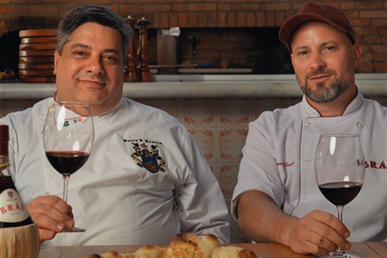 Dois homens sentados segurando uma taça de vinho com uma pizza em frente a eles