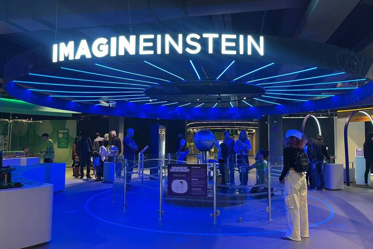 A imagem mostra uma exposição interativa chamada 'IMAGINEINSTEIN', com um grande letreiro iluminado no topo. O ambiente é predominantemente azul, com luzes neon e várias pessoas explorando diferentes estações interativas. No centro, há uma esfera metálica cercada por uma barreira de vidro. Ao fundo, há mais visitantes e outras áreas de exposição visíveis.