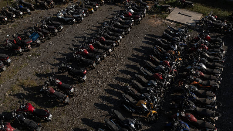 Imagem aérea de um pátio ao ar livre com várias motos estacionadas em fileiras organizadas. O chão é de cascalho e há uma área de concreto no canto superior direito. As motos são de diferentes modelos e cores, principalmente em tons escuros, com algumas em vermelho e amarelo.