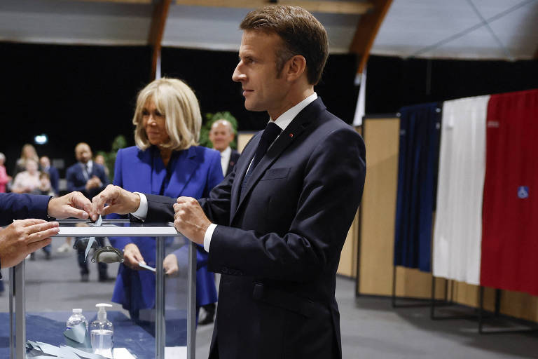 Macron, com terno preto, está depositando um voto em uma urna transparente. Ao fundo está a primeira-dama, uma mulher de cabelo loiro e com um casaco azul. Há cabines de votação com cortinas cujas cores (azul, branco e vermelho) formam a bandeira da França ao fundo