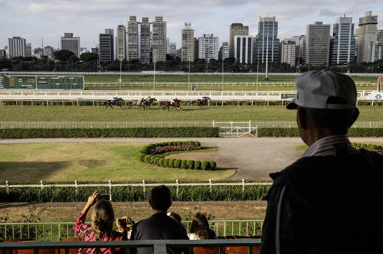 Público, fotografado de costas, acompanha um dos páreos das arquibancadas do Jockey Club; ao fundo aparece cavalos com jóqueis correndo na pista