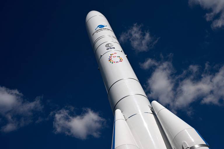 Imagem de um foguete Ariane 6 apontando para o céu, com um fundo de céu azul e algumas nuvens. O foguete é branco e possui vários logotipos, incluindo o da Arianespace, ESA (Agência Espacial Europeia) e outros símbolos coloridos.