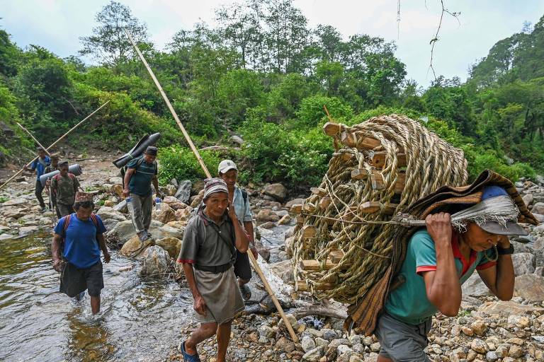 Um grupo de pessoas caminha por uma trilha rochosa e atravessa um riacho raso em uma área florestal. Eles carregam escadas de corda, feitas de materiais naturais, possivelmente bambu, nas costas. Alguns utilizam bastões de madeira para auxiliar na caminhada. A vegetação ao redor é densa e verde, indicando uma área de floresta tropical.