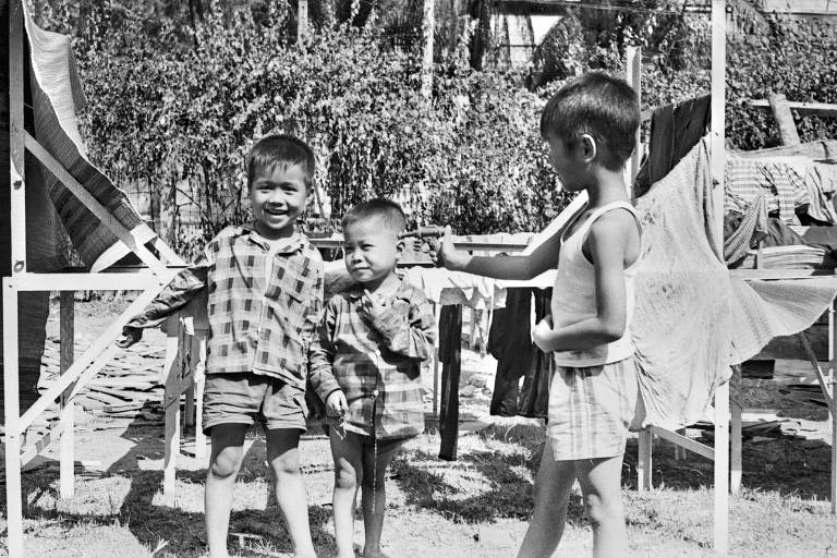 A imagem em preto e branco mostra três crianças brincando ao ar livre. Duas delas estão vestindo camisas de flanela xadrez e shorts, enquanto a terceira criança está usando uma camiseta sem mangas e shorts listrados. Ao fundo, há várias roupas penduradas em varais e uma vegetação densa.