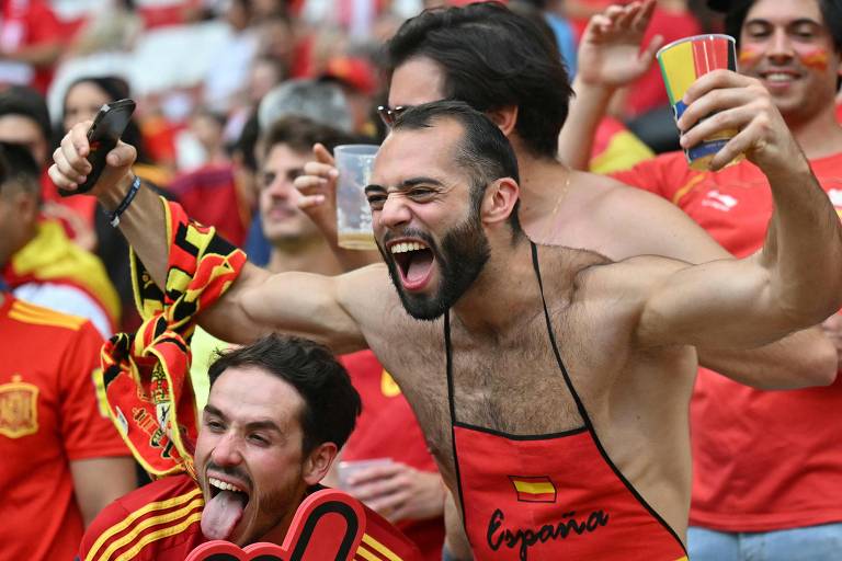 A imagem mostra um grupo de torcedores espanhóis comemorando em um estádio. Um homem no centro está sem camisa, usando um avental vermelho com a bandeira da Espanha e a palavra 'España' escrita. Ele está gritando e segurando um copo de bebida. Outro homem ao seu lado está fazendo uma careta com a língua para fora, vestindo uma camisa vermelha da seleção espanhola. Ao fundo, outros torcedores também estão comemorando, vestindo roupas nas cores da Espanha.