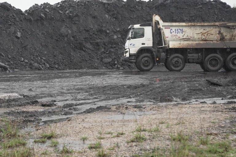 A imagem mostra um caminhão de carga estacionado em frente a uma grande pilha de carvão. O solo ao redor está molhado e há algumas áreas de vegetação rasteira. O caminhão tem uma placa com o número 'C-1578'.
