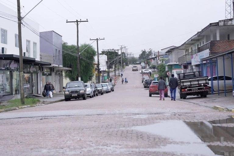 A imagem mostra uma rua residencial em um dia nublado. Há vários carros estacionados ao longo da rua e algumas pessoas caminhando. As casas e estabelecimentos comerciais estão alinhados em ambos os lados da rua. A rua é pavimentada com paralelepípedos e há postes de eletricidade ao longo dela.
