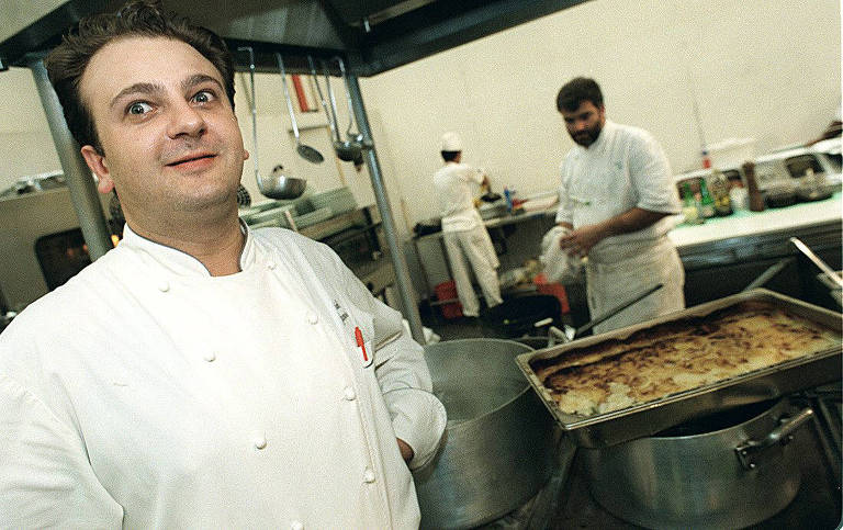 A imagem mostra dois cozinheiros em uma cozinha profissional. O cozinheiro em primeiro plano está usando um uniforme branco e parece estar sorrindo. Atrás dele, há outro cozinheiro também vestido de branco, que está preparando algo na bancada. Em primeiro plano, há uma grande assadeira com um prato gratinado. A cozinha está equipada com utensílios pendurados e várias superfícies de trabalho.