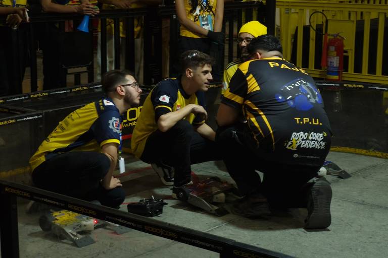 A imagem mostra três pessoas vestindo uniformes de equipe de robótica, ajoelhadas e concentradas em um robô no chão. Eles estão em um ambiente de competição, cercados por grades de proteção e outras pessoas ao fundo. Os uniformes são predominantemente amarelos e pretos, com logotipos e inscrições nas costas.