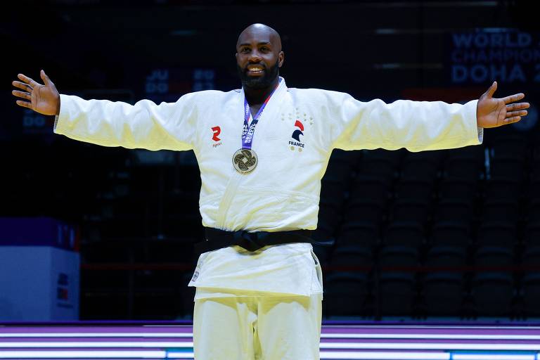 O judoca posa de braços abertos com uma medalha de ouro no peito e uma faixa preta na cintura.