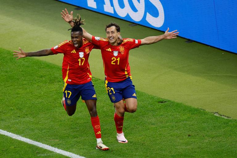 Dois jogadores de futebol, vestindo uniformes vermelhos com detalhes em amarelo e azul, comemoram com os braços abertos em um campo de futebol. O jogador à esquerda usa a camisa número 17 e o jogador à direita usa a camisa número 21. Ao fundo, há uma placa publicitária azul.